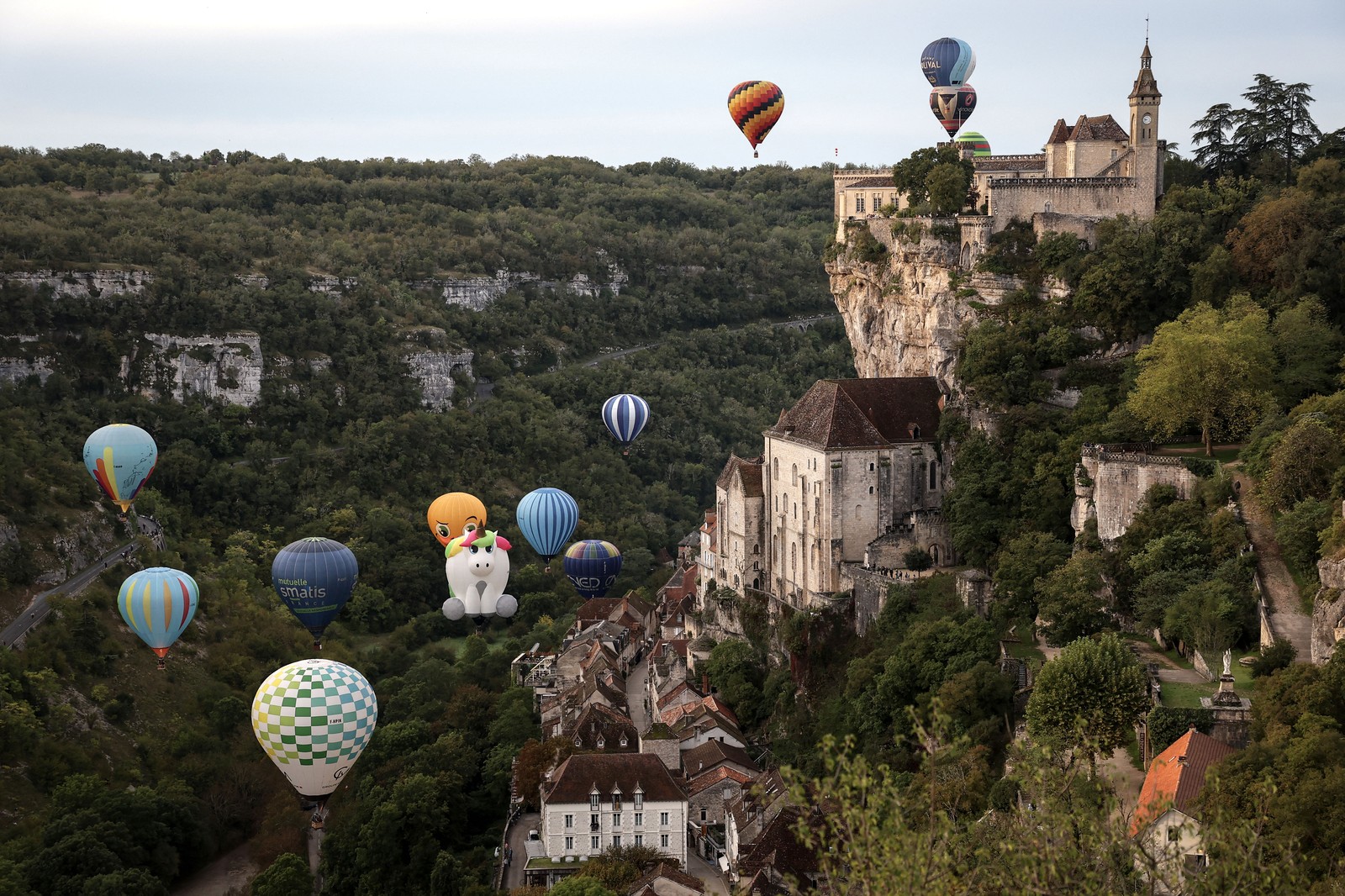 A dozen hot air balloons float along past historic cliffside structures in France.