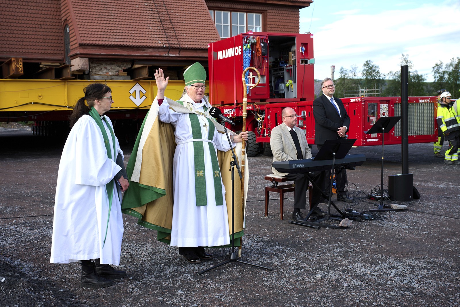 A bishop raises an arm during a blessing, beside a large church ready to be moved.