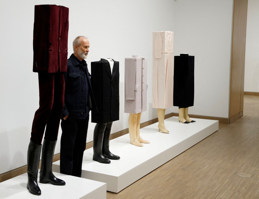 A person stands in an exhibition space beside five mannequins clothed in unusual boxy garments.