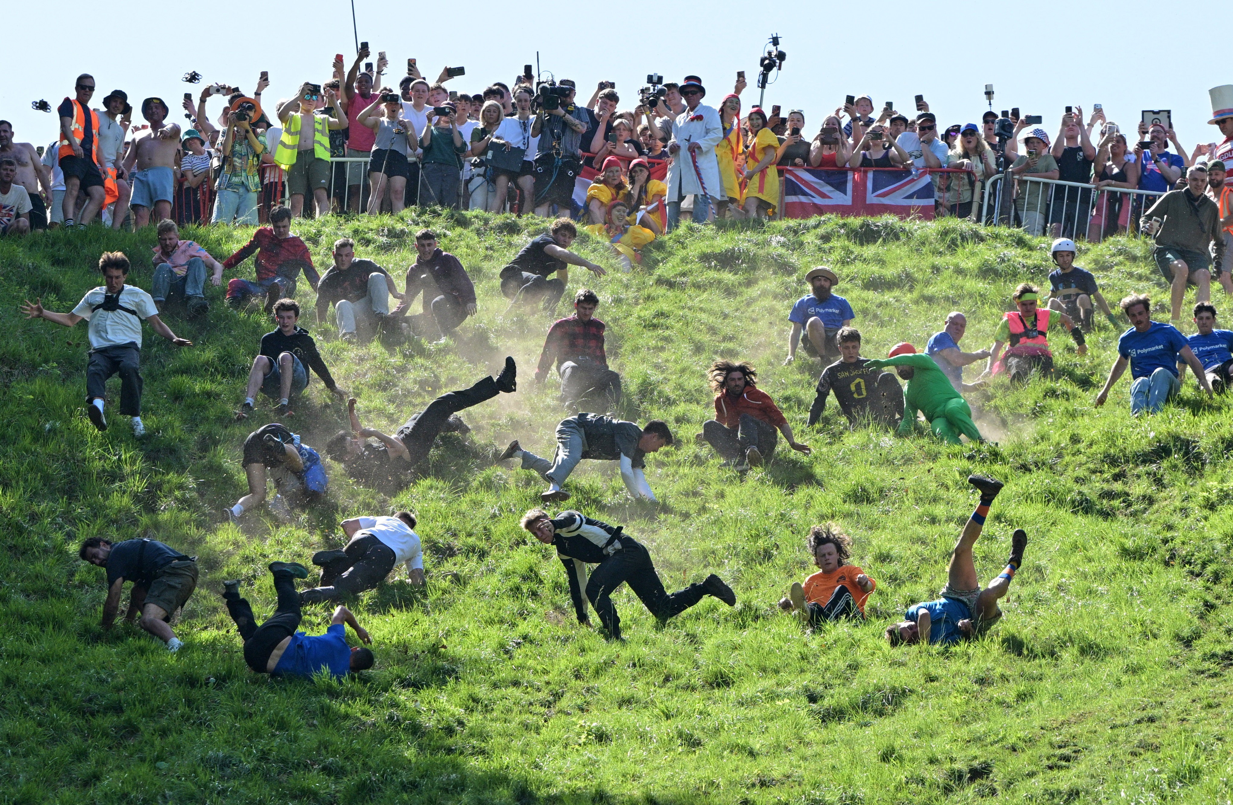Dozens of runners scoot and tumble down a steep grassy hill during a race, chasing a wheel of cheese.