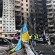 A photo shows bomb-damaged city buildings with emergency workers, burned cars, and a muddy Ukrainian flag in the foreground.
