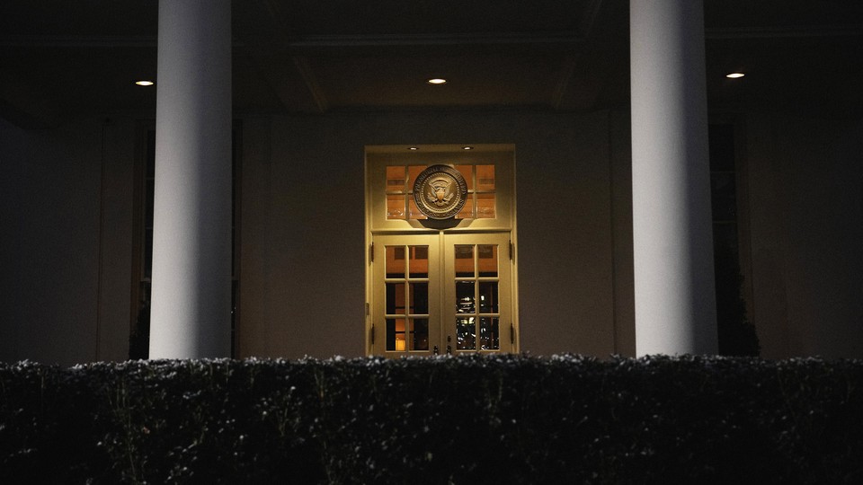 A color photograph of a White House door at night, seen behind a hedge and between two columns.