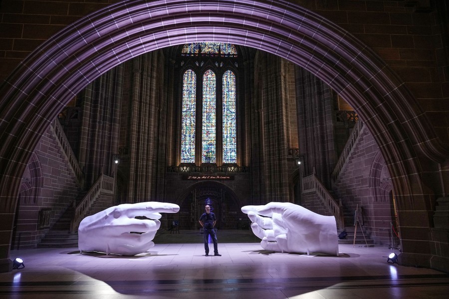 An artist stands in an open space in a cathedral, flanked by two large hand sculptures.
