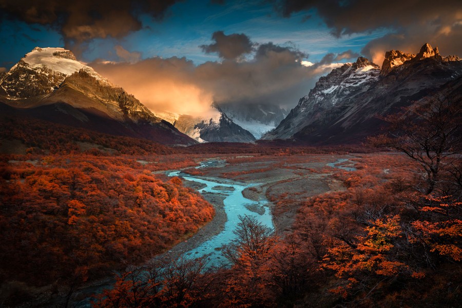 A landscape photograph of tall mountains and a river valley covered in autumn-colored bushes and trees.