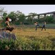 A girl sits on a rock blowing a gum bubble, while two girls in the distance play fight on a green field under a highway.