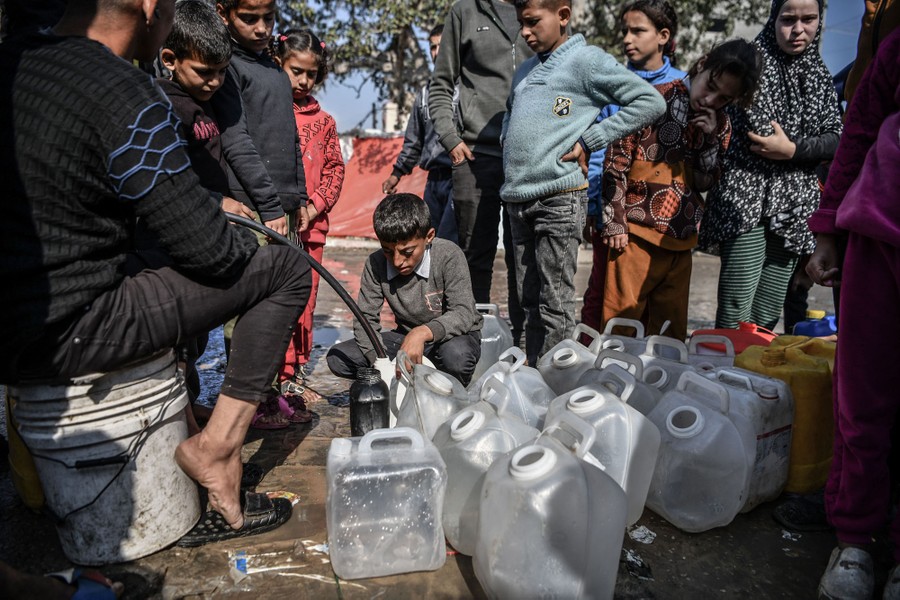 Children stand in a line, waiting beside water jugs that are being filled by a person with a hose.
