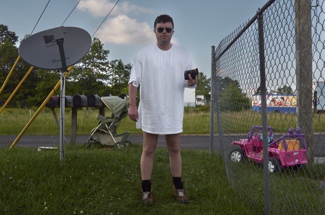 A man stands in a yard wearing sunglasses, no pants,  black socks , and shoes. Behind him is a satellite, pink toy car and a baby stroller.