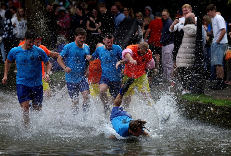 A crowd watches as several people compete for a football in a shallow riverbed, splashing and falling.