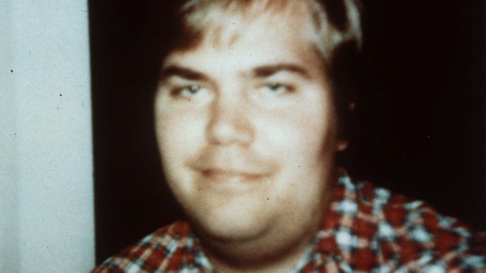 A photo of John Hinckley taken in 1999 shows him smiling and wearing a plaid shirt.