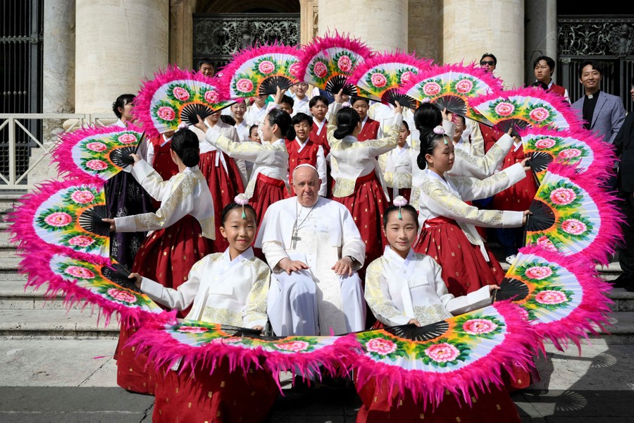 Pope Francis sits for a photo outside, surrounded by a ring of performers in costume, each holding decorative fans.