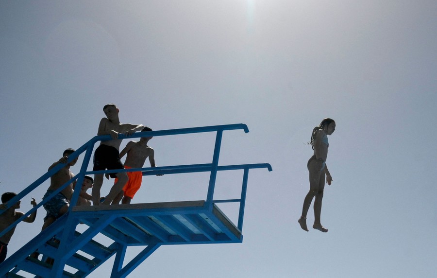 Several young people wait on a diving platform as another jumps off, appearing to hover in the air.