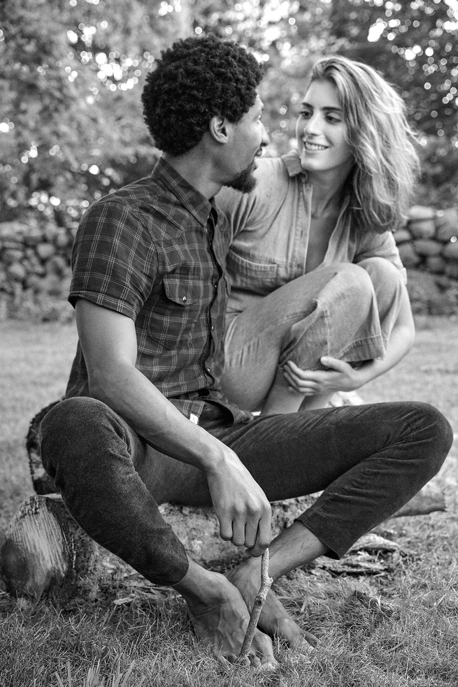 black-and-white photo of Jon sitting on log turned toward Suleika who is crouching next to him and smiilng