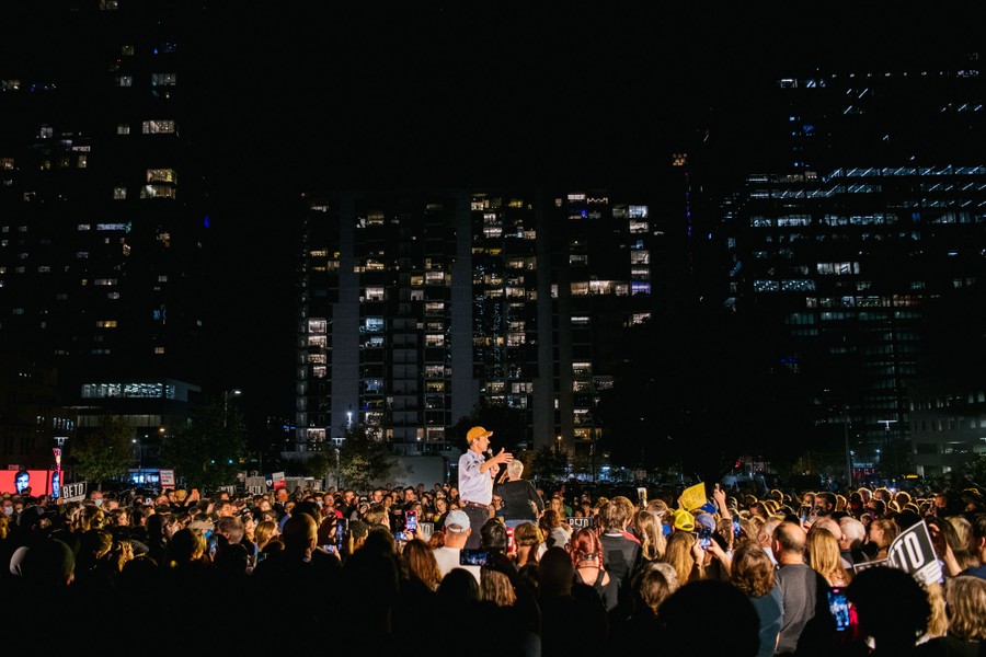 A large crowd of people gather around Beto O'Rourke as he speaks in a city square at night.