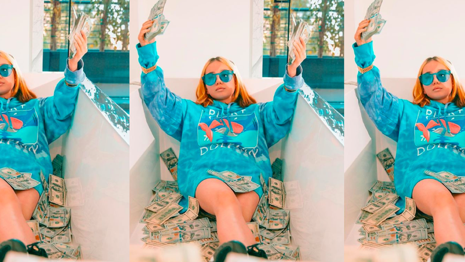 A young girl wearing a blue hoodie and blue sunglasses sitting in a bathtub full of dollar bills