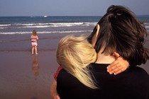 A woman holding a child and standing near another child, all by the ocean