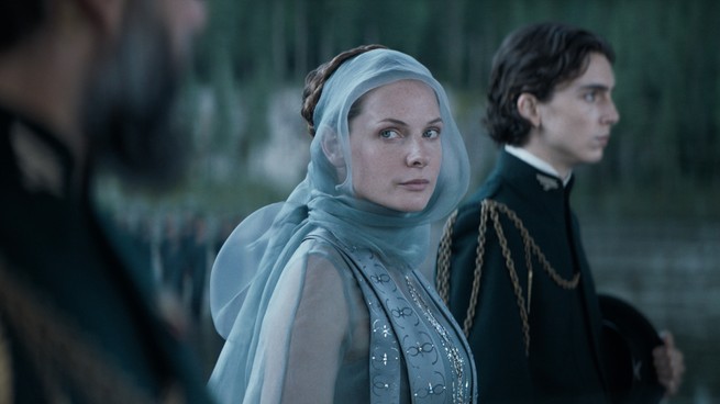 Rebecca Ferguson turns to look at the camera as she stands beside Timothée Chalamet, wearing a blue head scarf and blue shirt