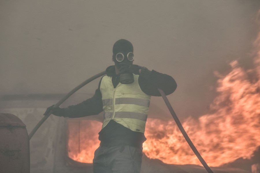 A person wearing a gas mask and a high-visibility safety vest carries a hose near a wildfire.