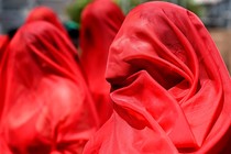 Demonstrators stand together, wearing bright-red fabric draped over their heads.