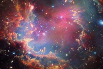 A star cluster is shown inside a large nebula of multicolored gas and dust. The material forms dark ridges and peaks of gas and dust surrounding the cluster, lit on the inner side, while layers of diffuse, translucent clouds blanket them.