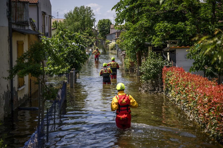 Several rescue workers walk along a road through thigh-deep floodwater.