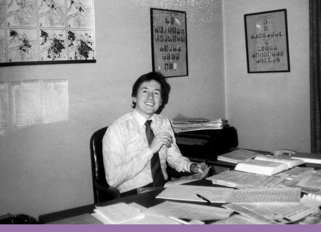 a man smiles sitting at an office desk with littered with papers