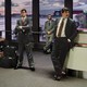 Photo of some suited men standing around an airport waiting area looking bored
