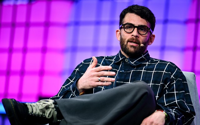 Photograph of Hasan Piker speaking with a microphone attached to his face. He is seated in a gray chair with his legs crossed.