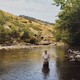 Color photo of a man in waders and a plaid shirt standing knee-deep in a stream holding a fly-fishing rod with hills in the background.