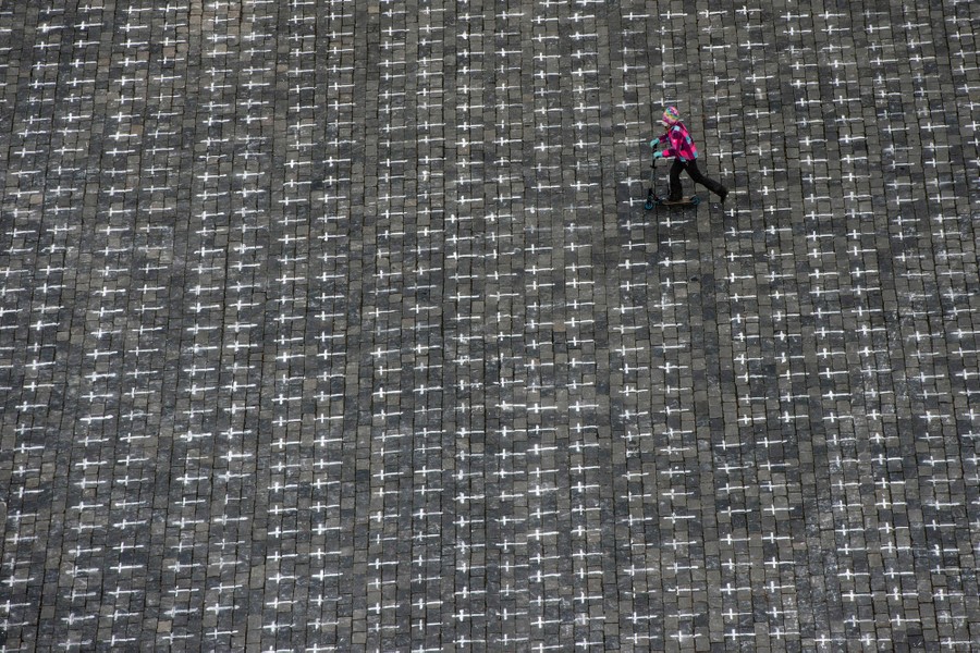 A child rides a scooter across a wide public square where hundreds of small crosses have been drawn.