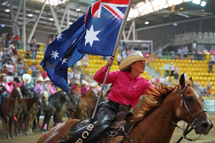 A woman rides a horse during a rodeo in an arena, carrying an Australian flag.
