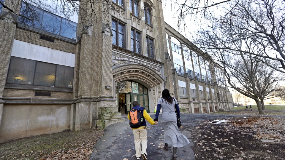 A mother and her son walk hand-in-hand into a brick school building. They are unrecognizable from the back.