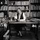 black-and-white photo of woman sitting behind desk next to sofa in home library in front of shelves filled with books