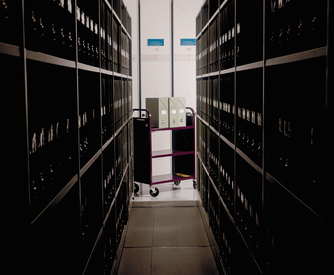 photo looking down an aisle of a library archive with rows of shelves and a rolling library cart at the end