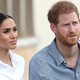 Meghan Markle, wearing a white button-up shirt with her hair slicked back into a low ponytail, and Prince Harry, wearing a chambray shirt, look off to the side of the camera.