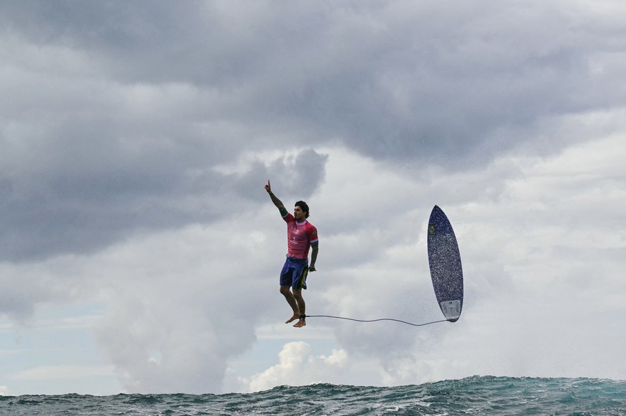 A surfer points upward while jumping above a wave with his surfboard beside him, appearing to stand casually upright in midair.