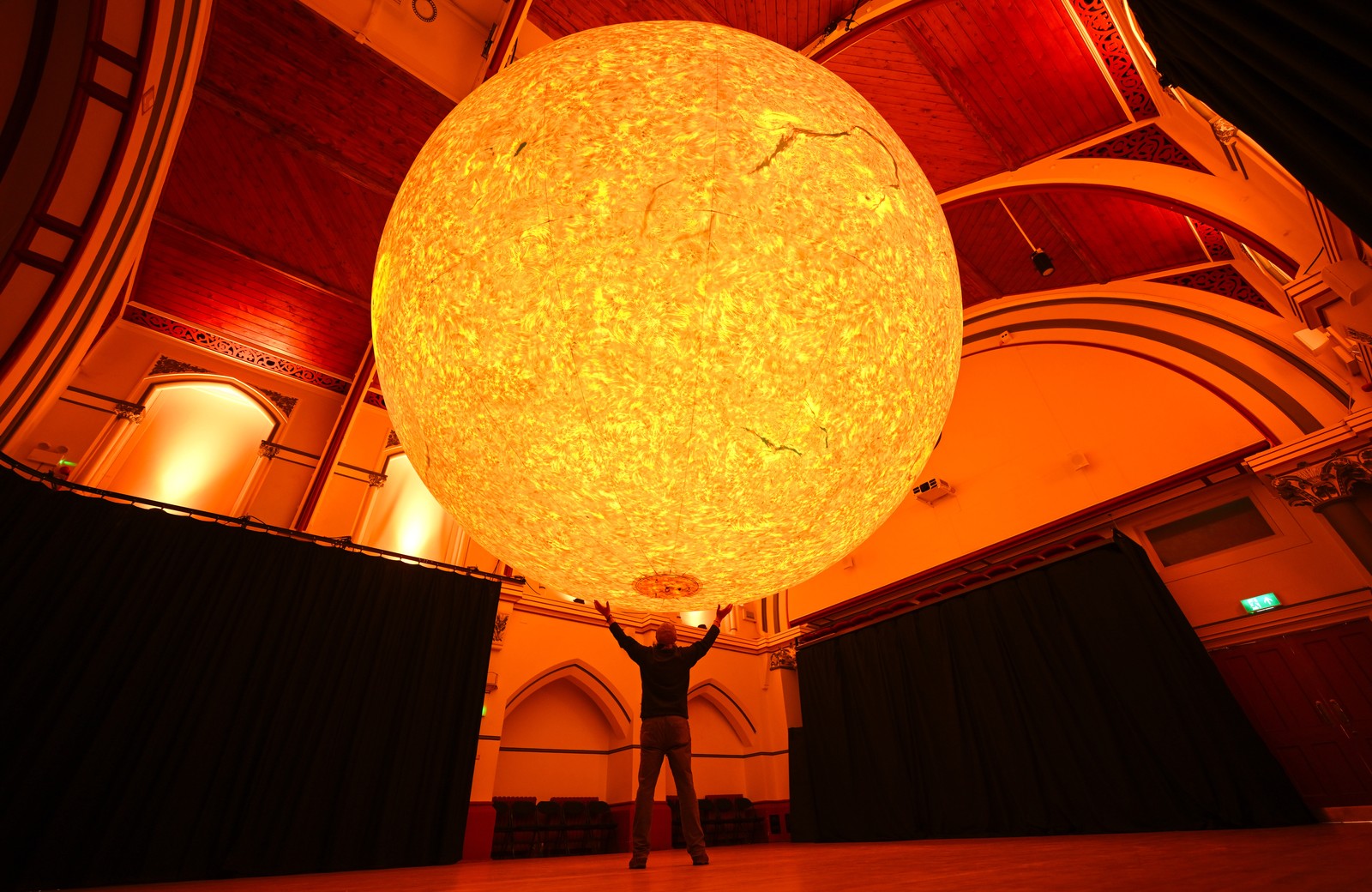 A person stands beneath a huge sculpture of the sun, suspended from a ceiling inside an exhibit space.
