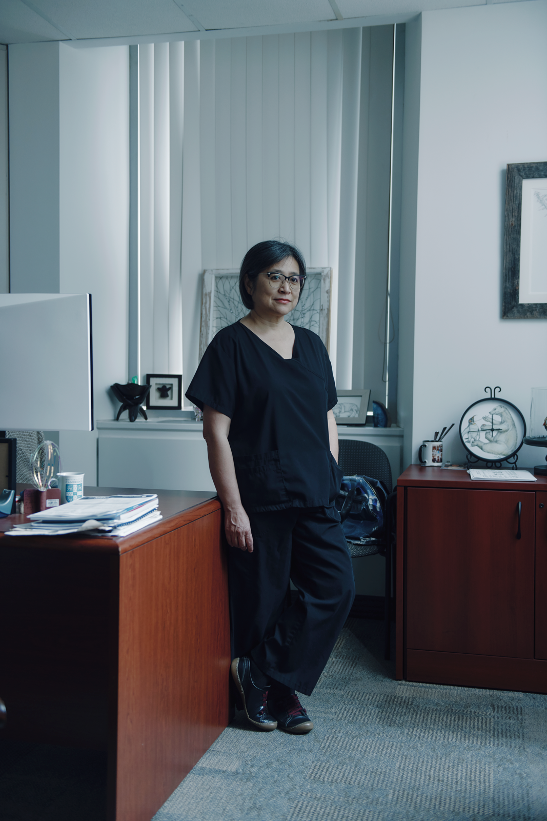 photo of woman with glasses and dark hair wearing black medical scrubs and standing near wooden desk by window