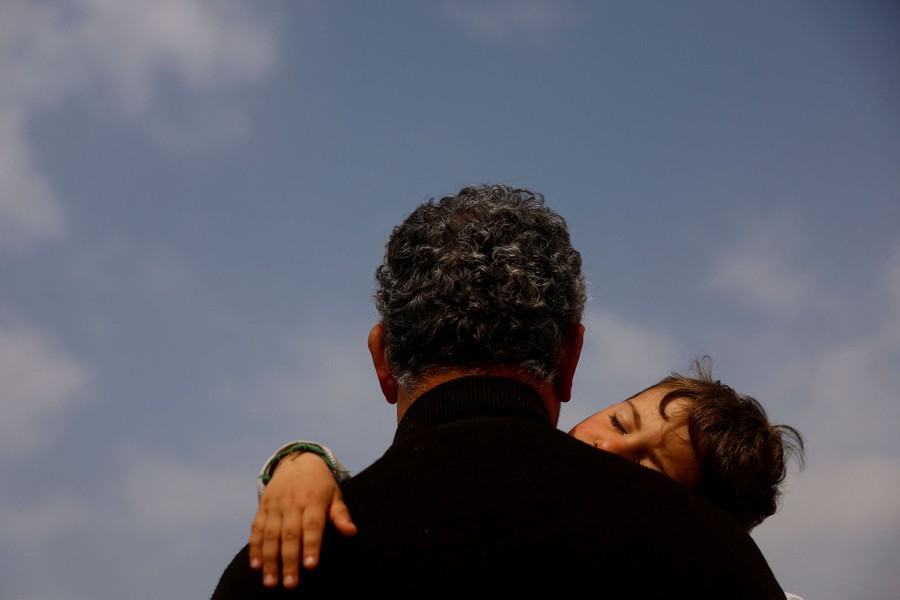 A man is seen from behind, carrying his young grandson, who snoozes on his shoulder.