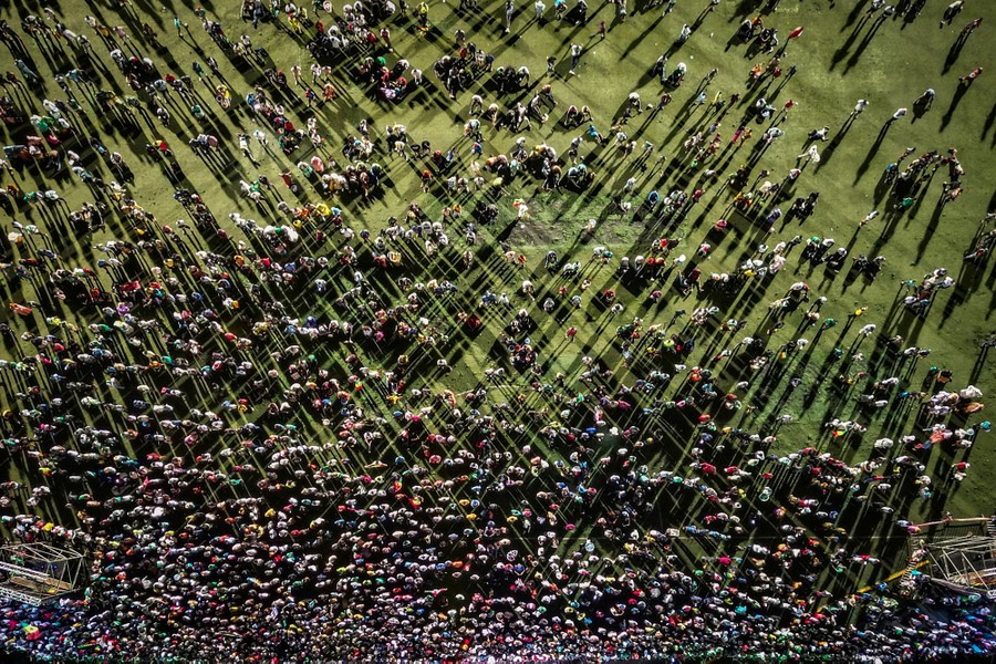 An overhead view of a large gathering crowd in a grassy field, lit from several sides, casting long, crisscrossing shadows