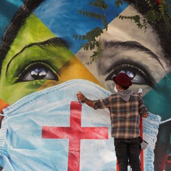 An artist stands in front of a large mural of the face of a young person wearing a mask.