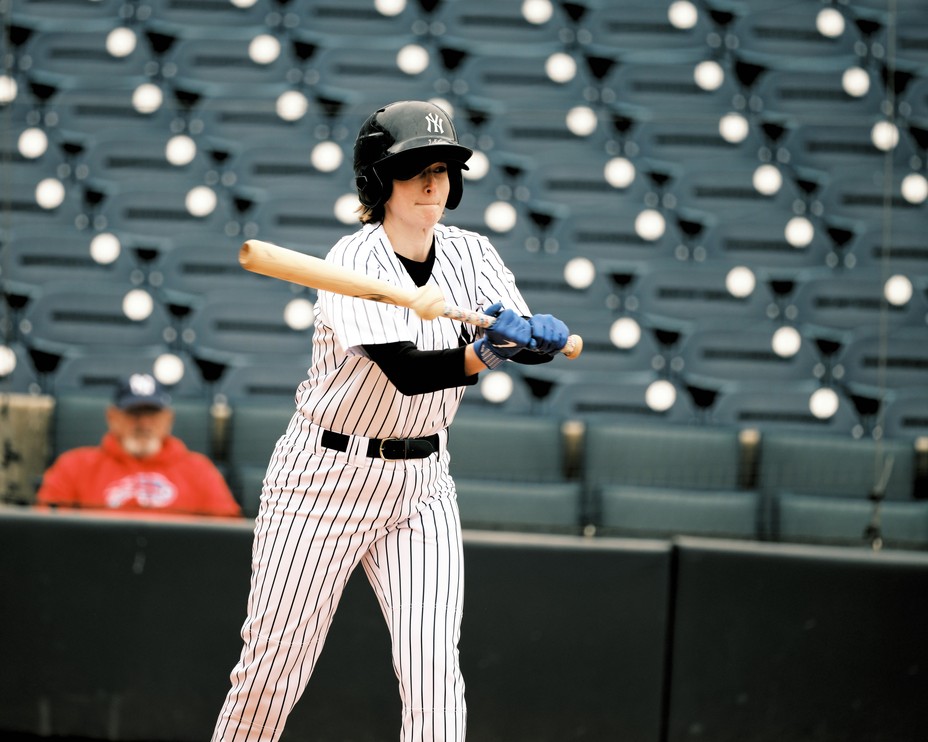 photo of woman in baseball uniform swinging bat at a baseball with mostly empty stadium seats behind her