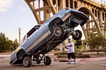 photo of hydraulic car resting on back wheels with front wheels in air above the head of man standing behind it, with tall arched bridge in background