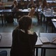 A student's back is photographed as she raises her hand. She wears her hair in long pigtail braids.