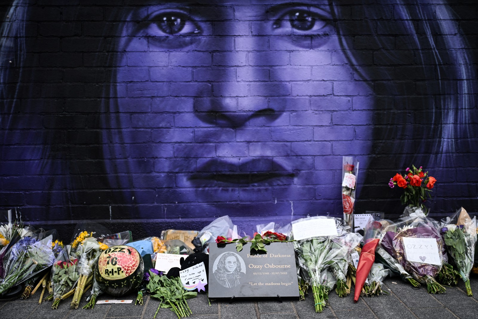 Flowers are laid on the ground beside a mural of Ozzy Osbourne painted on a wall.