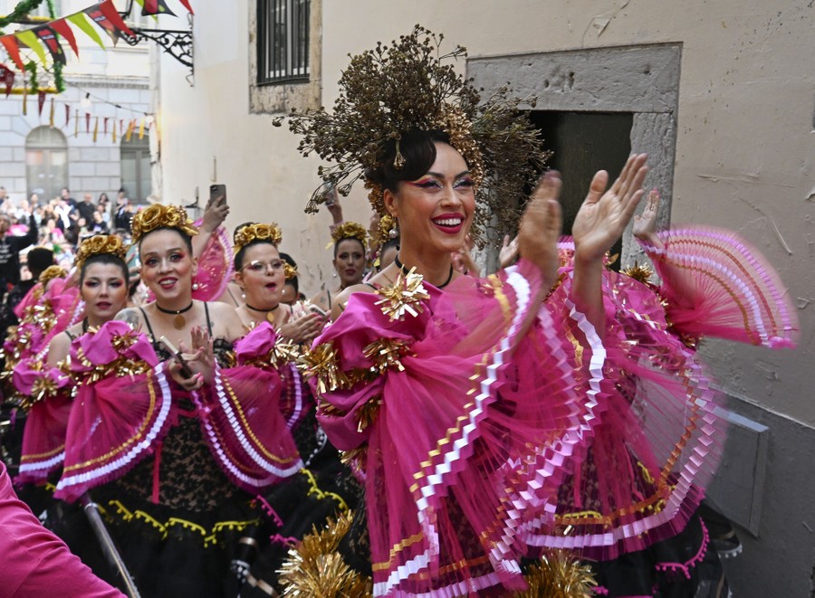 Costumed performers clap while walking through a narrow street.