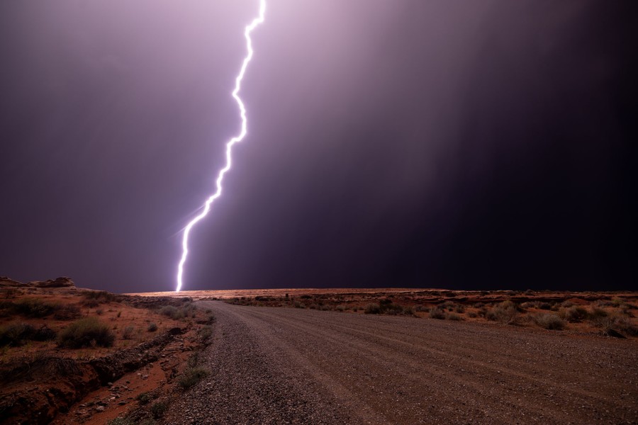 A single bolt of lightning in a dark sky illuminates arid ground beside a gravel road.
