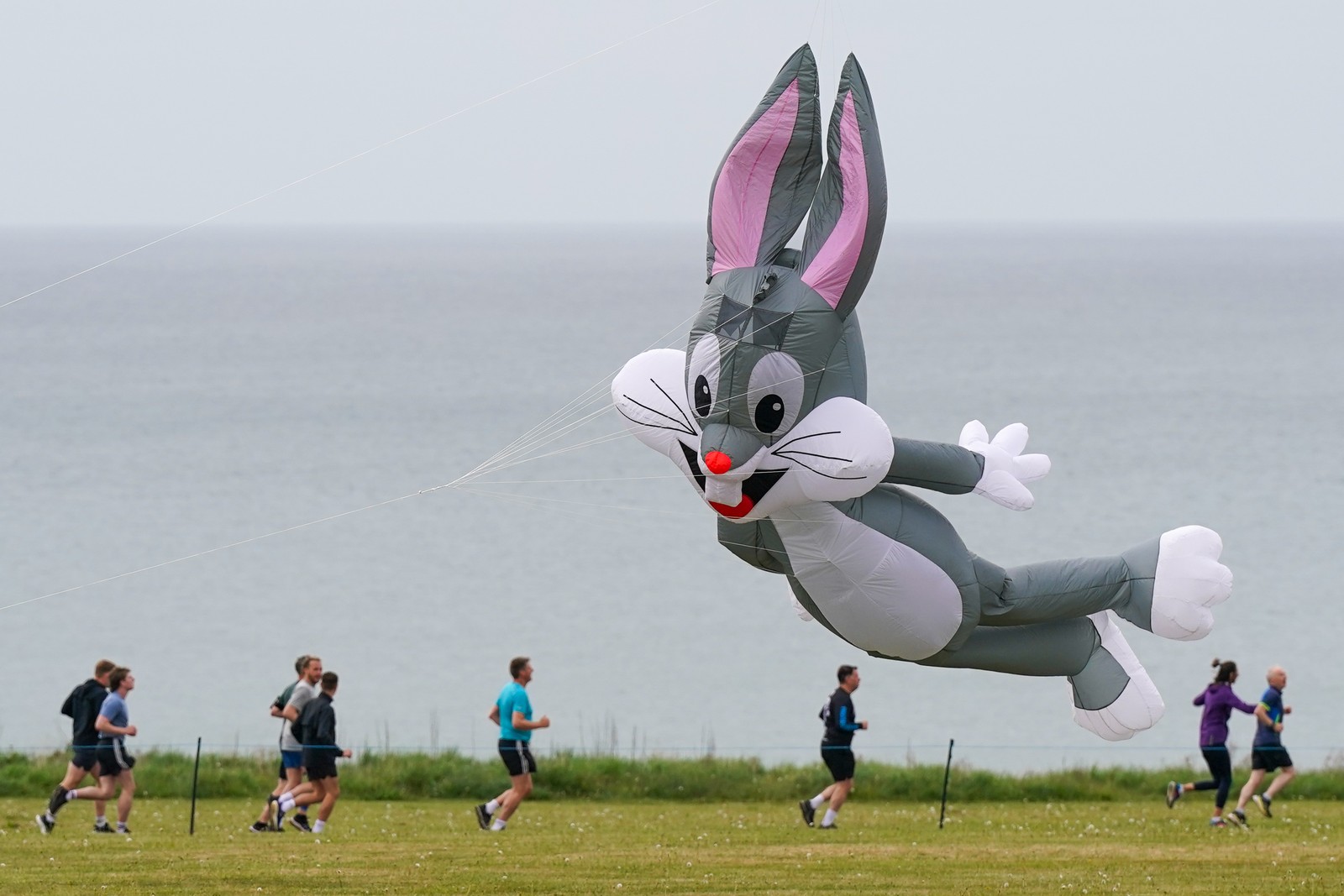 People run on a path beneath a large kite shaped like Bugs Bunny.