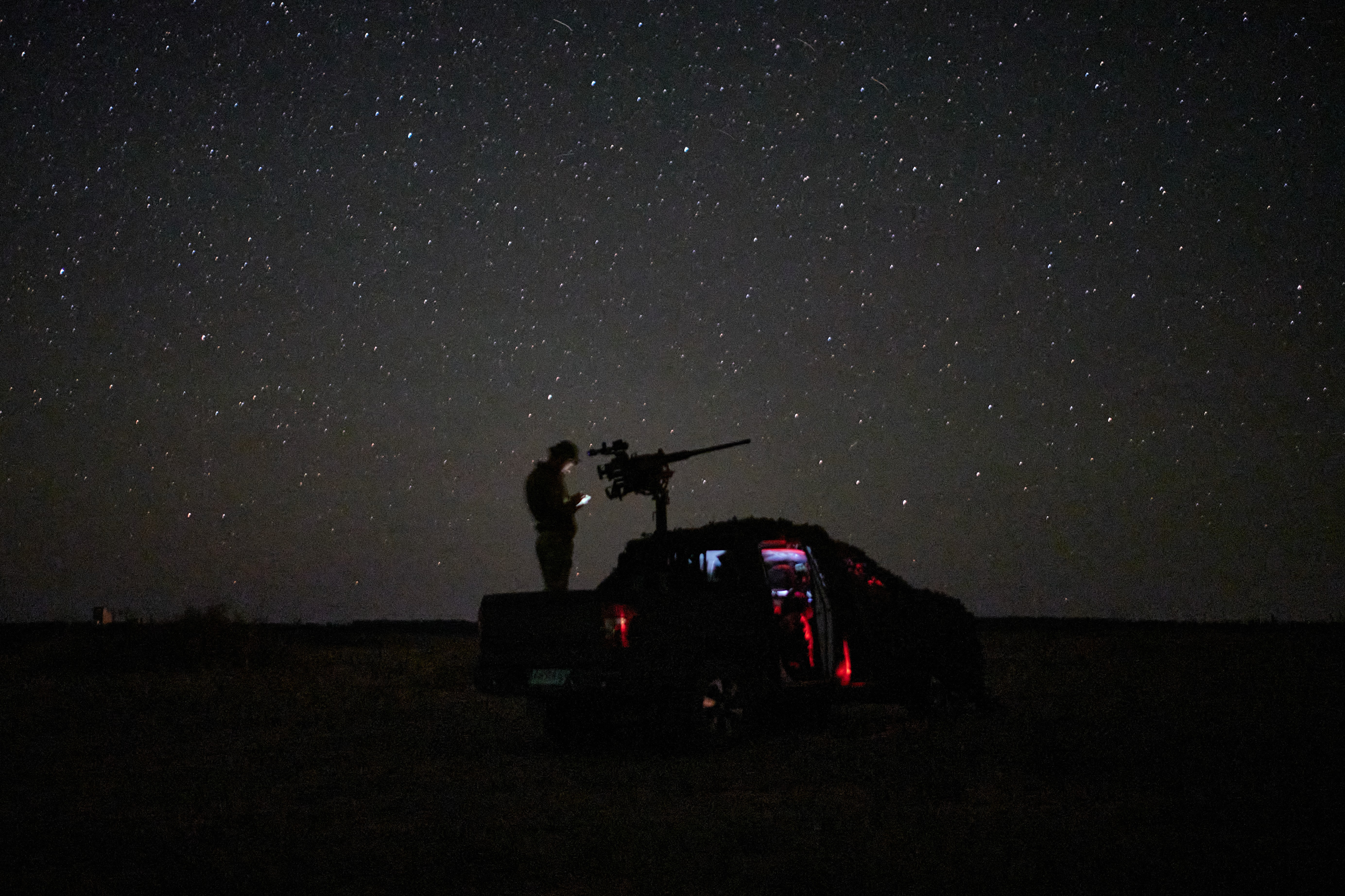 A soldier stands in a truck bed beside a mounted gun, seen under stars at night.