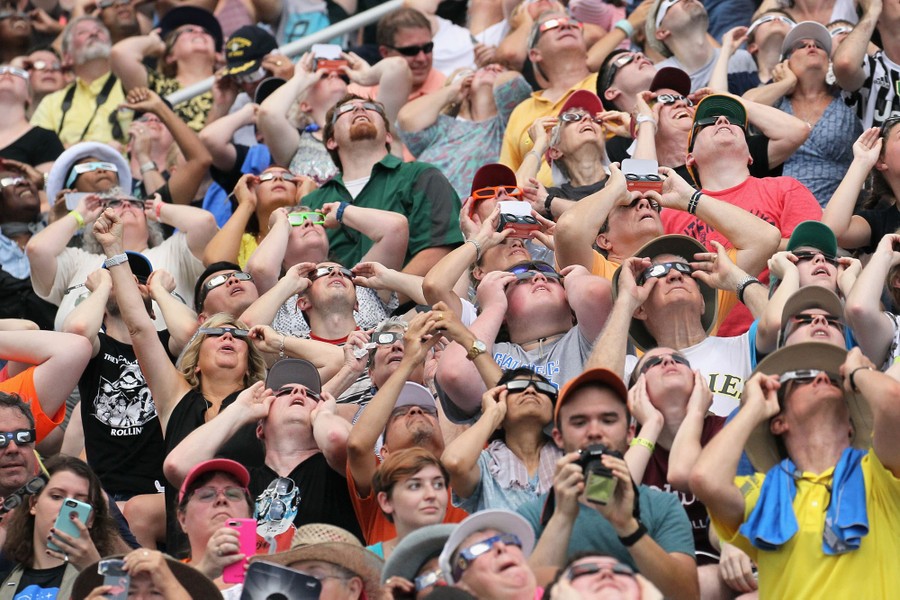 Many people in a stadium, most wearing eclipse glasses, look upward.