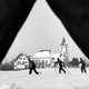 A black and white photograph of a winter scene of skiers, seen through the outstretched legs of another skier